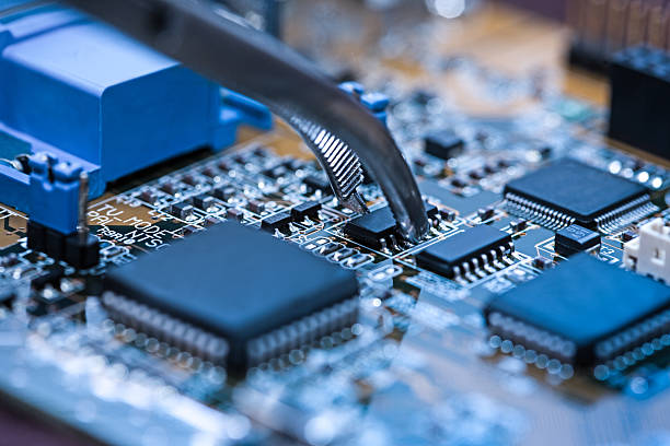 Close-up of tweezers holding transistor on blue circuit board.